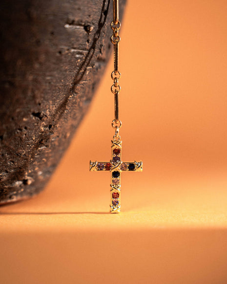 Gold cross earring with colored gemstones on a textured surface