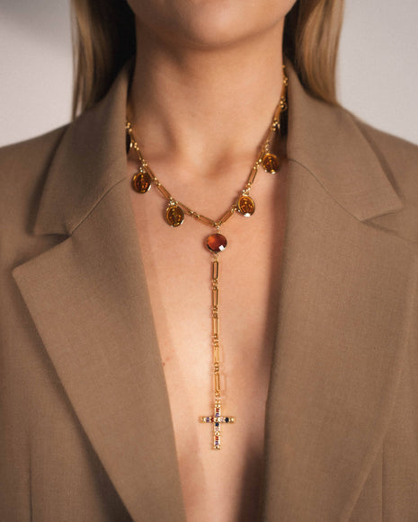 Gold necklace with a cross pendant worn by a person in a brown blazer.
